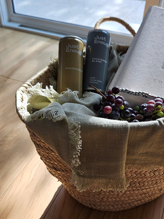 A woven basket with “Just Enough” Chardonnay and Sauvignon Blanc wine cans, a book, and grapes, arranged for a relaxed picnic scene.