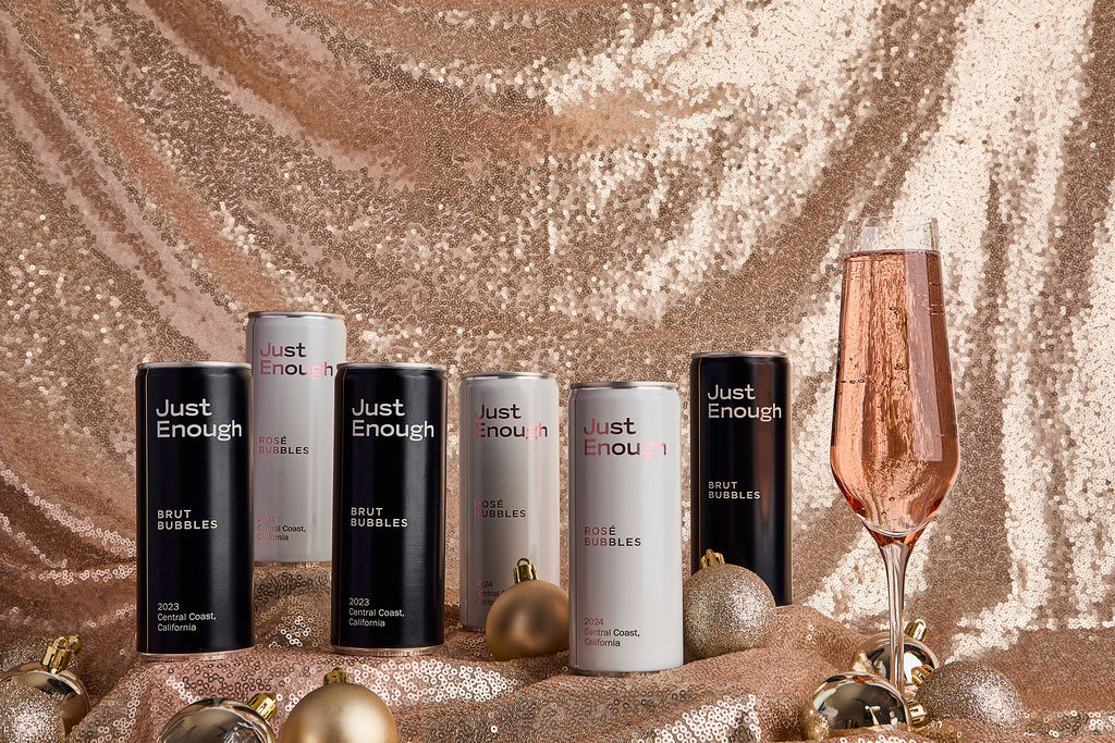 Just Enough Wines Brut and Rosé Bubbles cans with a champagne glass on a festive gold backdrop.