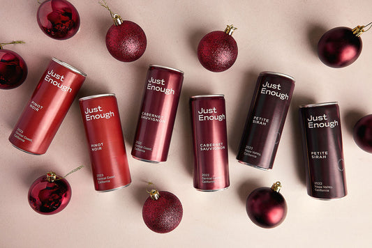 Holiday display of Just Enough Wines red varietals in single-serve cans surrounded by festive red ornaments.
