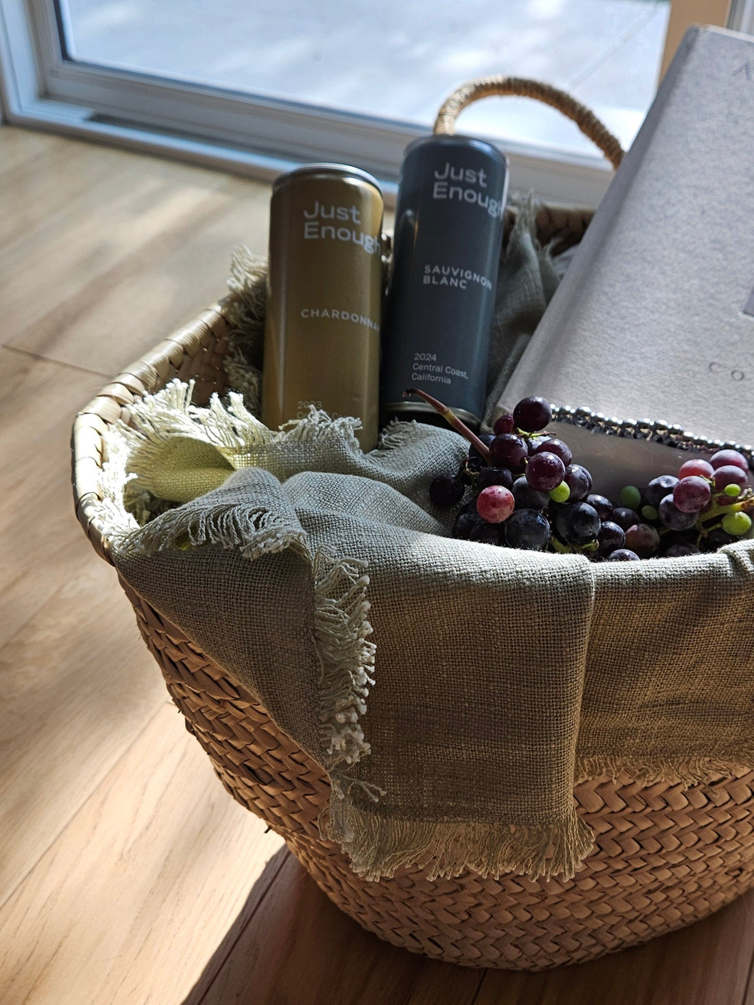 A woven basket with “Just Enough” Chardonnay and Sauvignon Blanc wine cans, a book, and grapes, arranged for a relaxed picnic scene.