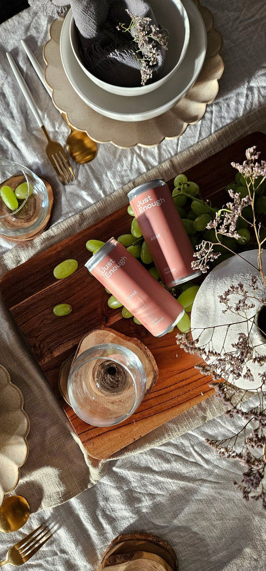 Two cans of Just Enough Wines Rosé placed on a table beside grapes and a decorative vase.