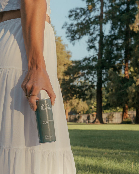 A woman holding a can of Just Enough, Sauvignon Blanc in an open garden.