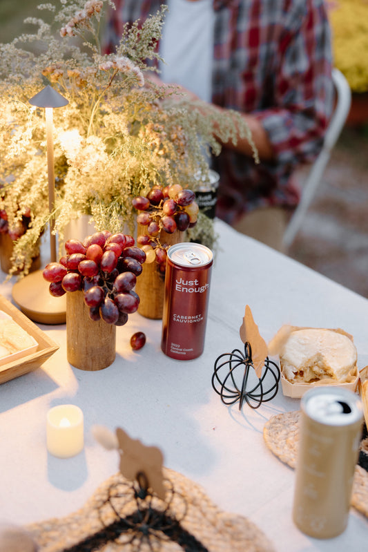 An outdoor table setup featuring a can of Just Enough Wines Cabernet Sauvignon surrounded by grapes, artisan cheese, and festive Halloween decor.