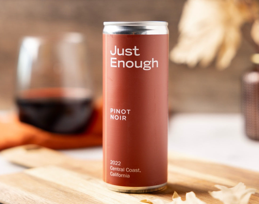 A can of Just Enough Pinot Noir from the Central Coast, California, is placed on a wooden board beside a glass of red wine.