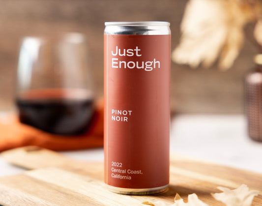 A can of Just Enough Pinot Noir from the Central Coast, California, is placed on a wooden board beside a glass of red wine.