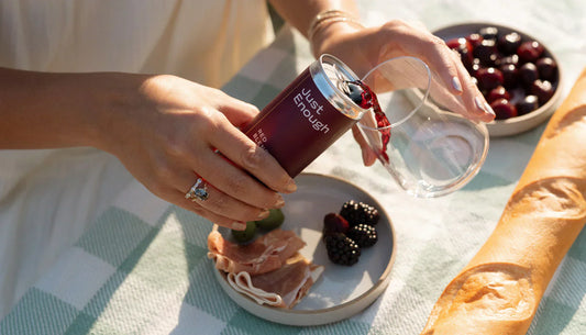 Person pours Just Enough Red Blend into a glass during a picnic with blackberries, prosciutto, and cherries.