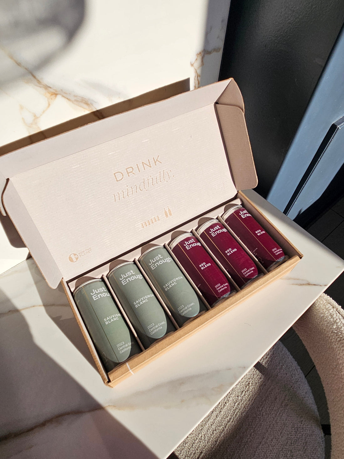 Box of six Just Enough Wines cans featuring Sauvignon Blanc and Red Blend placed on a chair.