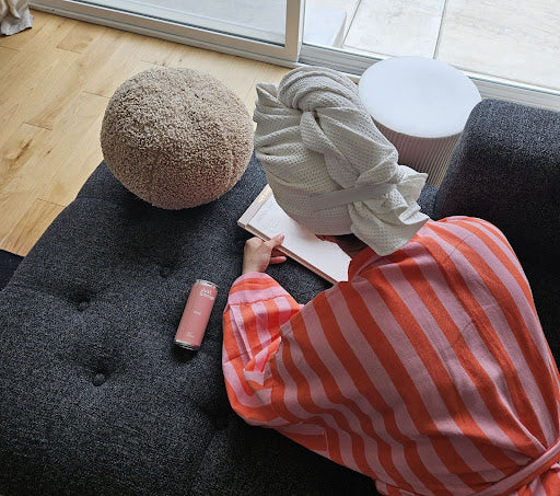 Woman relaxing on a sofa reading a book with a Just Enough rosé wine can beside her in a calm, cozy home setting.