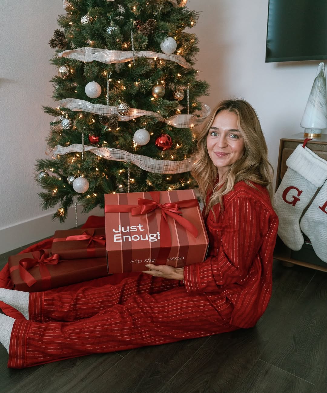 Woman in pajamas sitting in front of a Christmas tree, holding Just Enough Wines’ 2025 Wine Advent Calendar in a festive gift box.